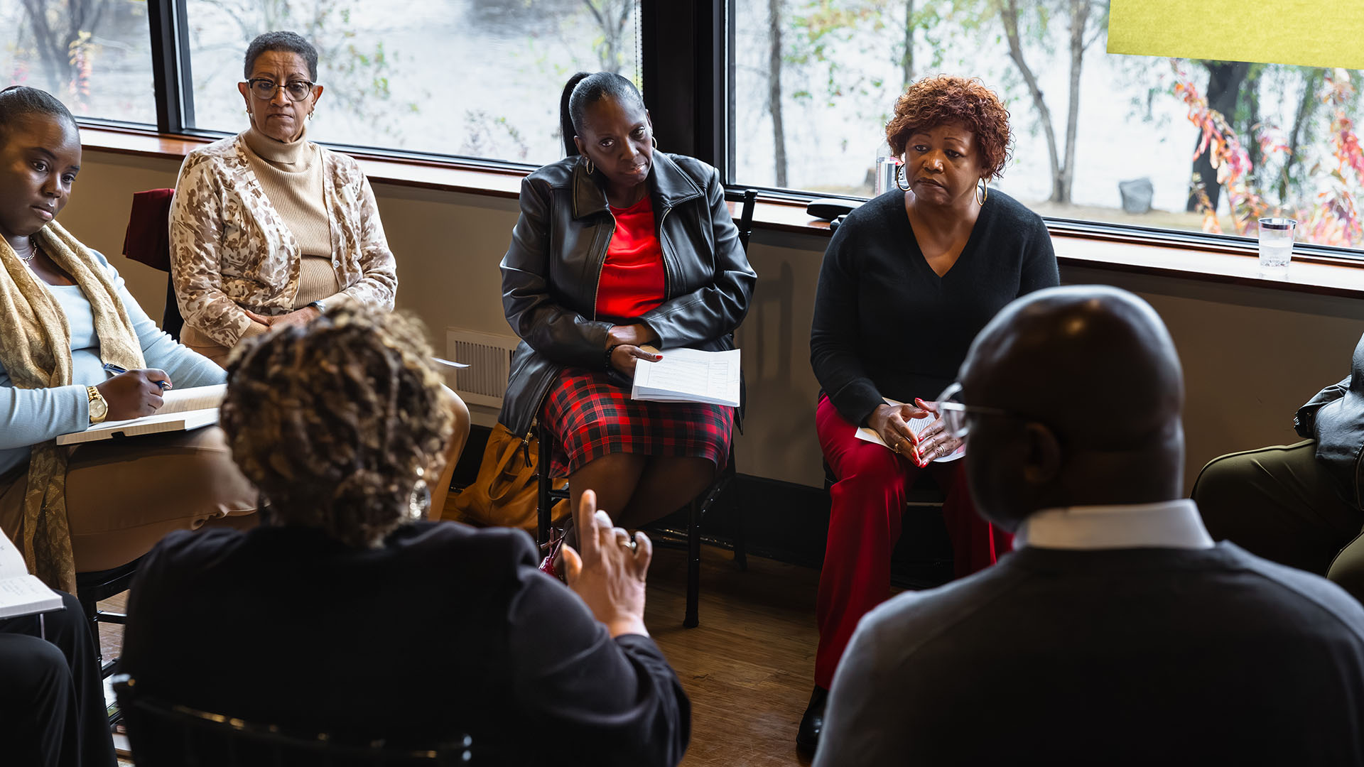 A diverse group of adults sit in a circle indoors, attentively listening and taking notes during a discussion. Large windows behind them reveal trees and a river outside.